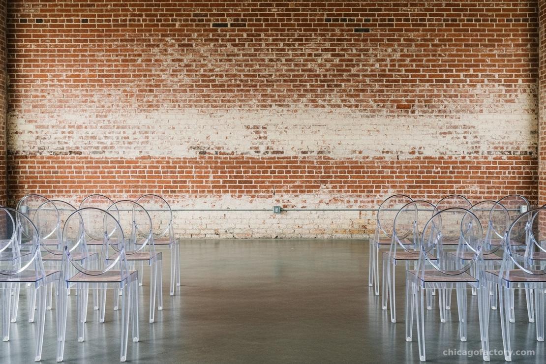 Clear ghost chairs lined up for a ceremony against an exposed brick wall, letting the texture show through.