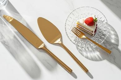 A modern gold cake cutting set resting on a marble table next to a slice of wedding cake.
